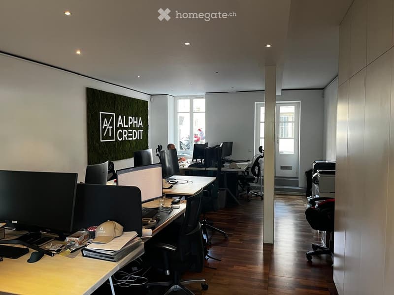 Office space with multiple desks, chairs, computers, and office supplies. Wooden flooring and large windows provide natural light.