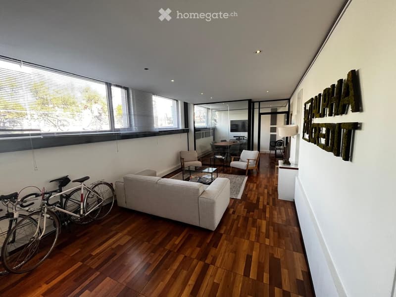 Modern living room with wooden flooring, large windows, sofa, coffee table, chairs, and a bicycle near the entrance.