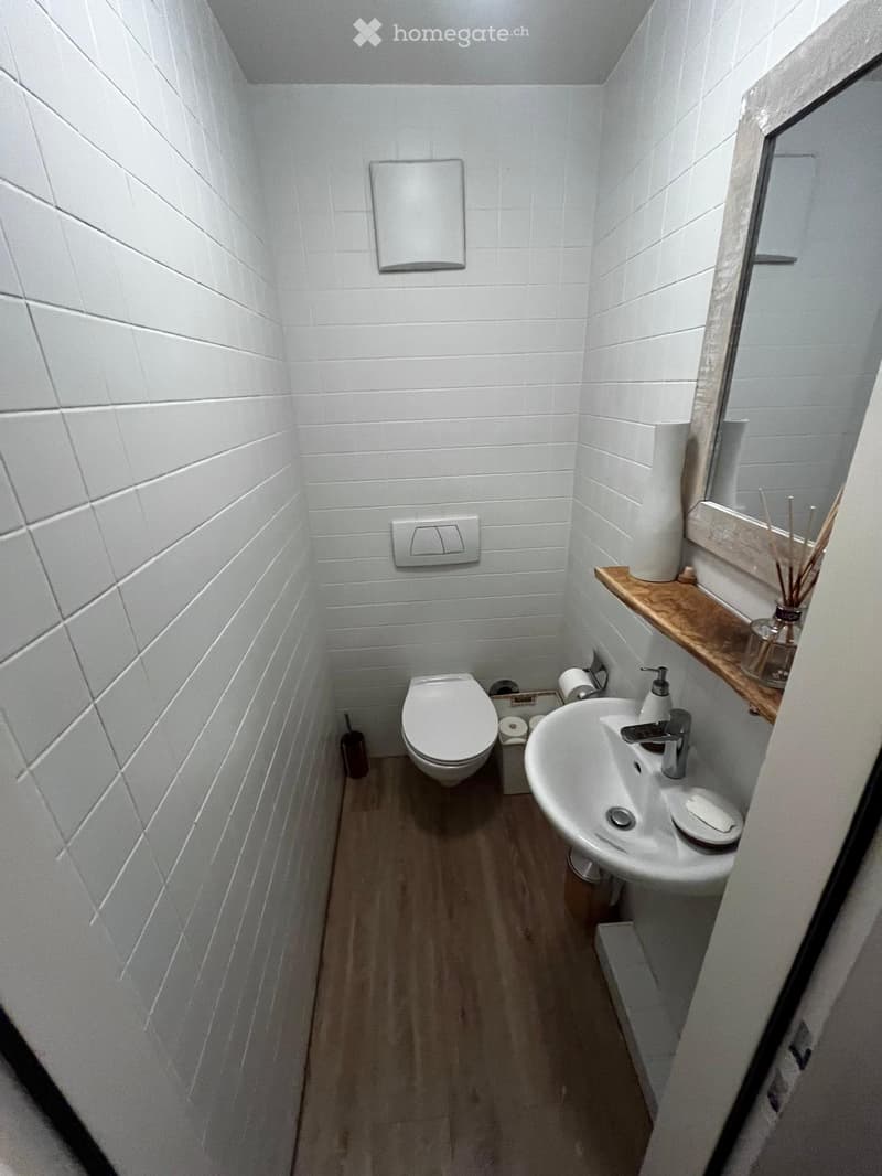 Modern, compact bathroom with white tiled walls, wooden floor, toilet, sink, and a small wooden shelf holding a mirror and decorative items.