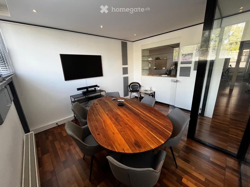 Modern, minimalist room with a round wooden table surrounded by six grey chairs, a TV screen mounted on the wall, a small black cabinet, and a white kitchen area with shelves and a stool.