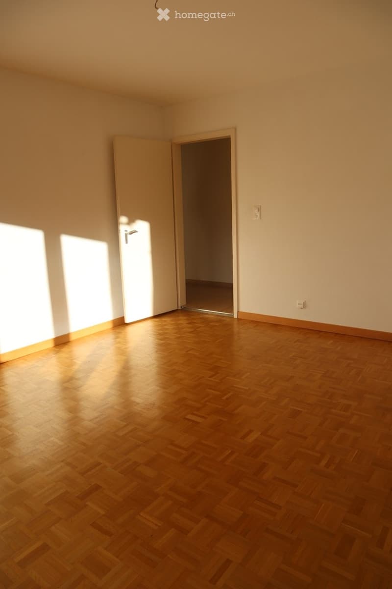 Empty room with wooden herringbone flooring, white walls, and a partially open door leading to another space.