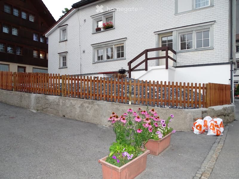 A raised garden area with potted flowers, a wooden fence, and a concrete retaining wall. The house in the background has white siding and multiple windows.