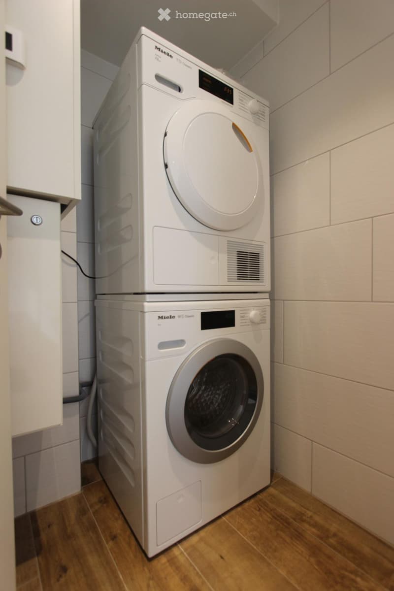 A compact bathroom with a stacked washing machine and dryer, white tiled walls, and wooden flooring.