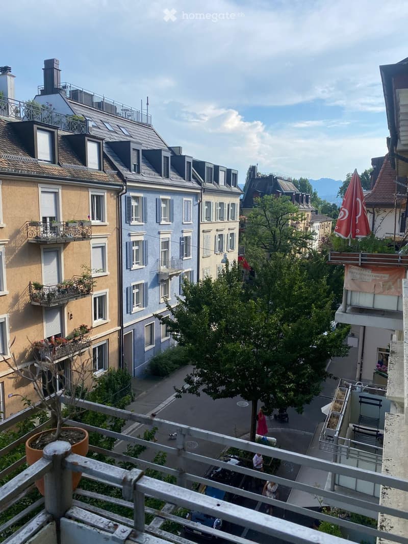 Balcony overlooking a narrow street lined with multi-story residential buildings, featuring balconies, shutters, and potted plants.