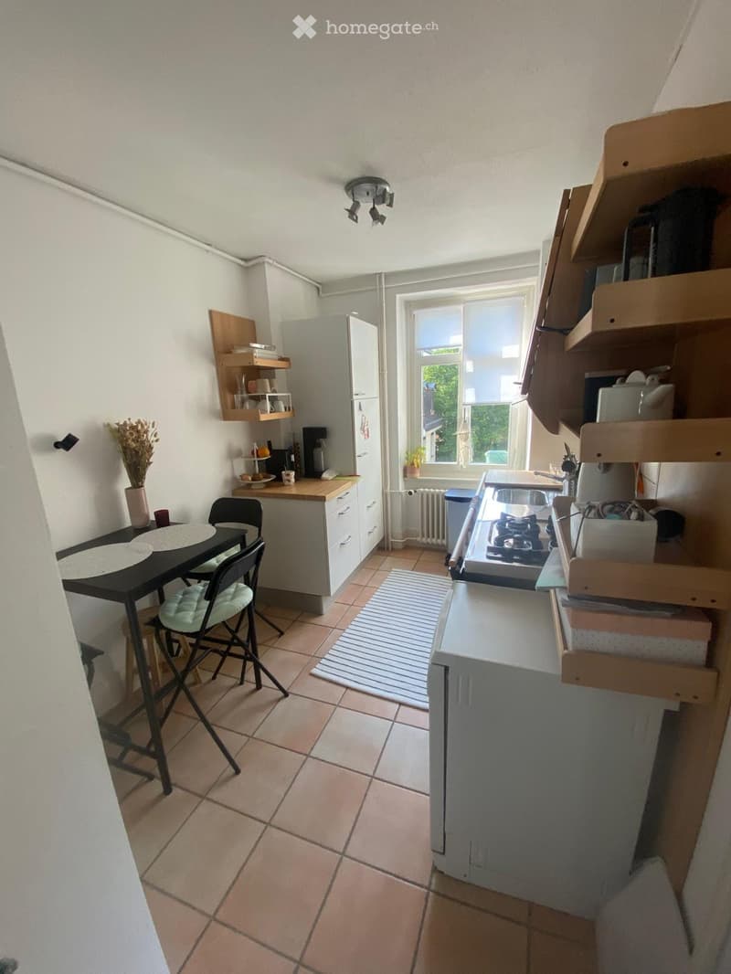 Compact kitchen with tiled floor, white walls, and wooden shelves. Includes a refrigerator, stove, sink, dining table with chairs, and various kitchen items on shelves and counters.