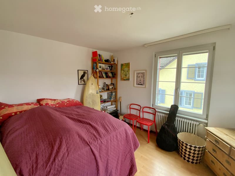 Cozy bedroom with a double bed covered in a purple blanket, red chairs, a guitar and case, a bookshelf with books and a surfboard, and framed pictures on the walls.