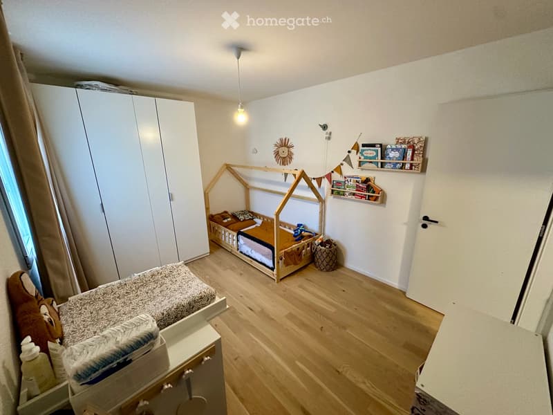 Cozy children's bedroom with wooden floor, two beds (one raised on a platform), a wardrobe, shelves with books and toys, and a basket on the floor.