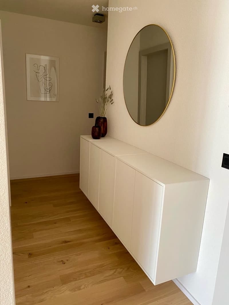 A modern hallway with light wooden flooring, white cabinetry, a large round mirror with a gold frame, and a framed abstract artwork on the wall.