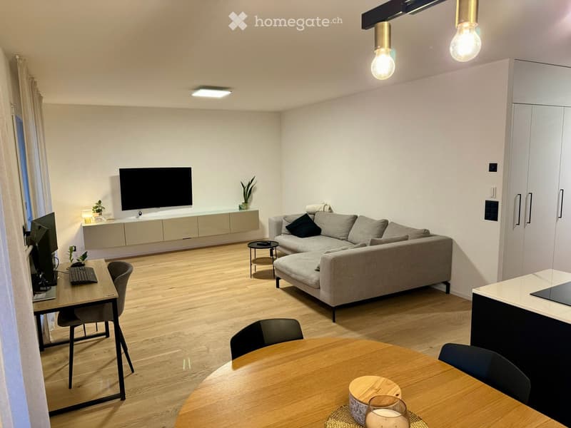 Modern living room with a grey sectional sofa, coffee table, dining table, laptop, and TV. Features wooden flooring, large windows, and built-in storage.