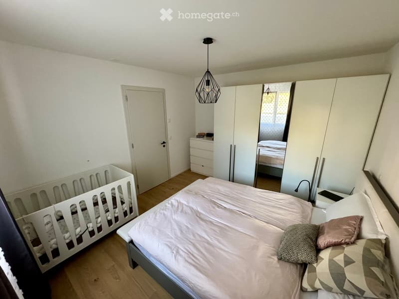 Bright bedroom with a double bed, pillows, a wardrobe with a mirror, a nightstand, and a baby crib. Wooden flooring and white walls.