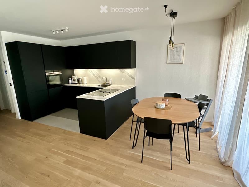 Modern kitchen with black cabinets, a central island, and a round dining table with chairs. The kitchen features a stove, sink, and coffee maker, with a picture frame on the wall.