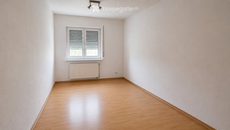 Empty room with light wooden flooring, white walls, a single window with blinds, and a radiator below it.