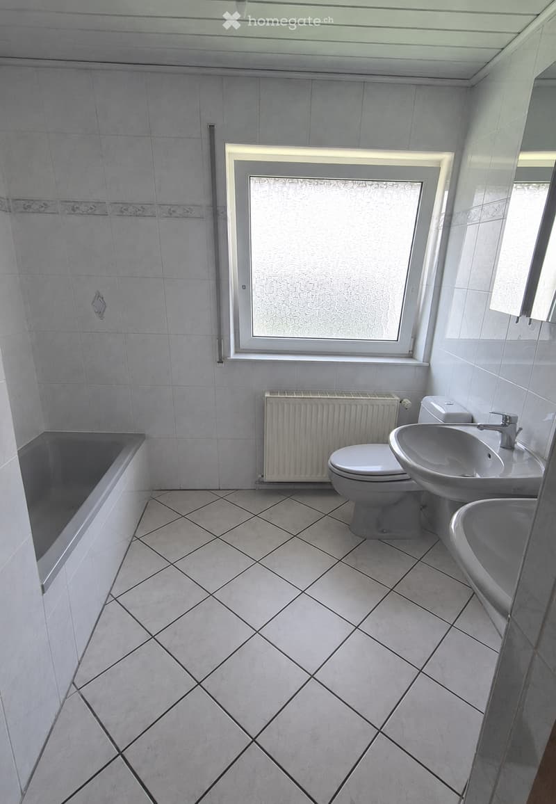 Bathroom with white tiled walls and floor, featuring a bathtub, toilet, sink, and radiator. Frosted window provides natural light.