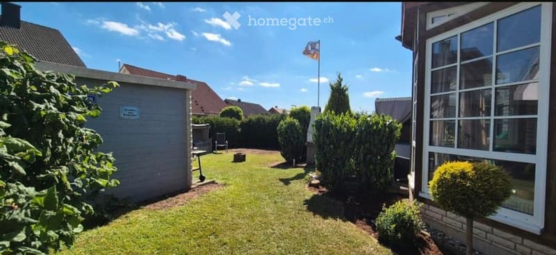 Well-maintained garden with green lawn, trimmed bushes, and a small seating area near the house.