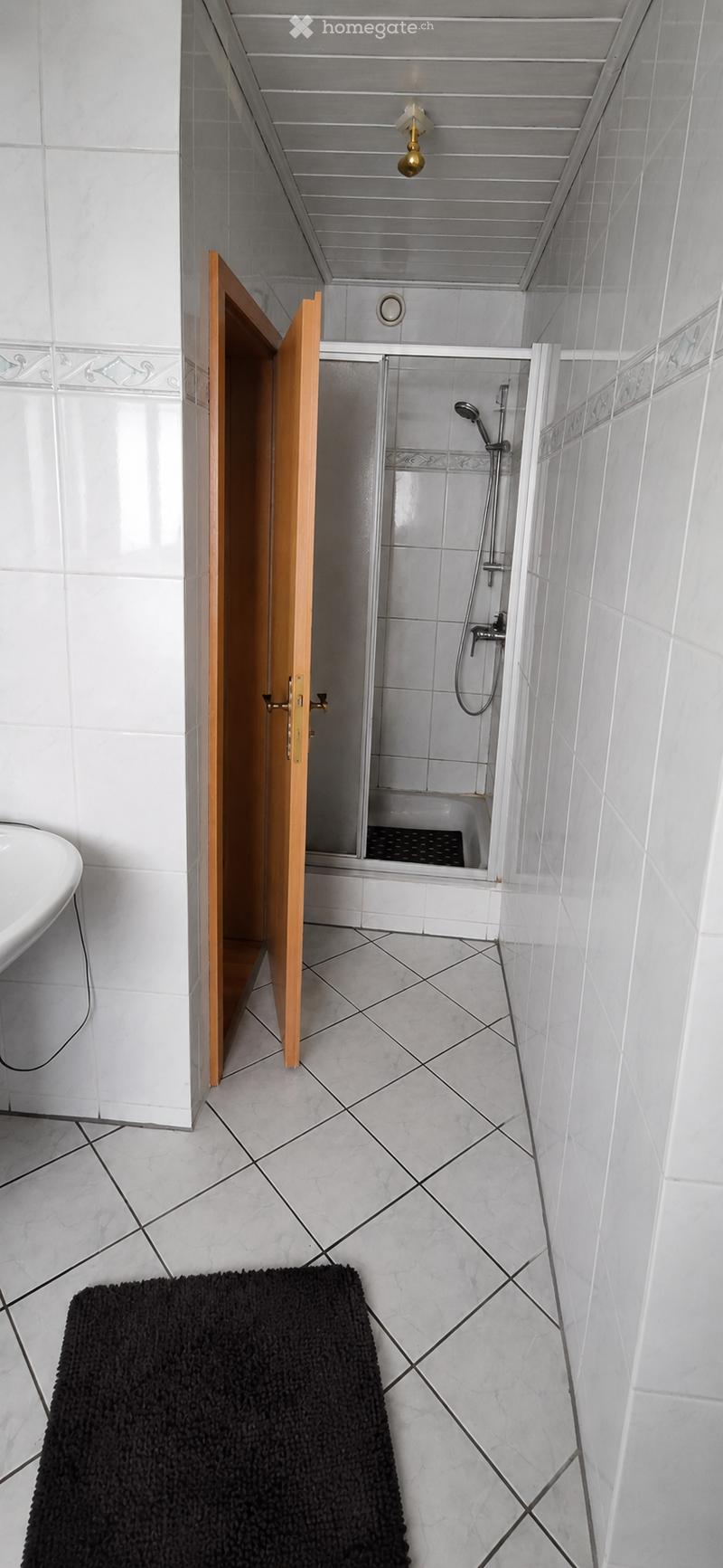 Modern bathroom with white tiles, a bathtub, a shower area with glass enclosure, a wooden door, and a black bathroom rug on the floor.