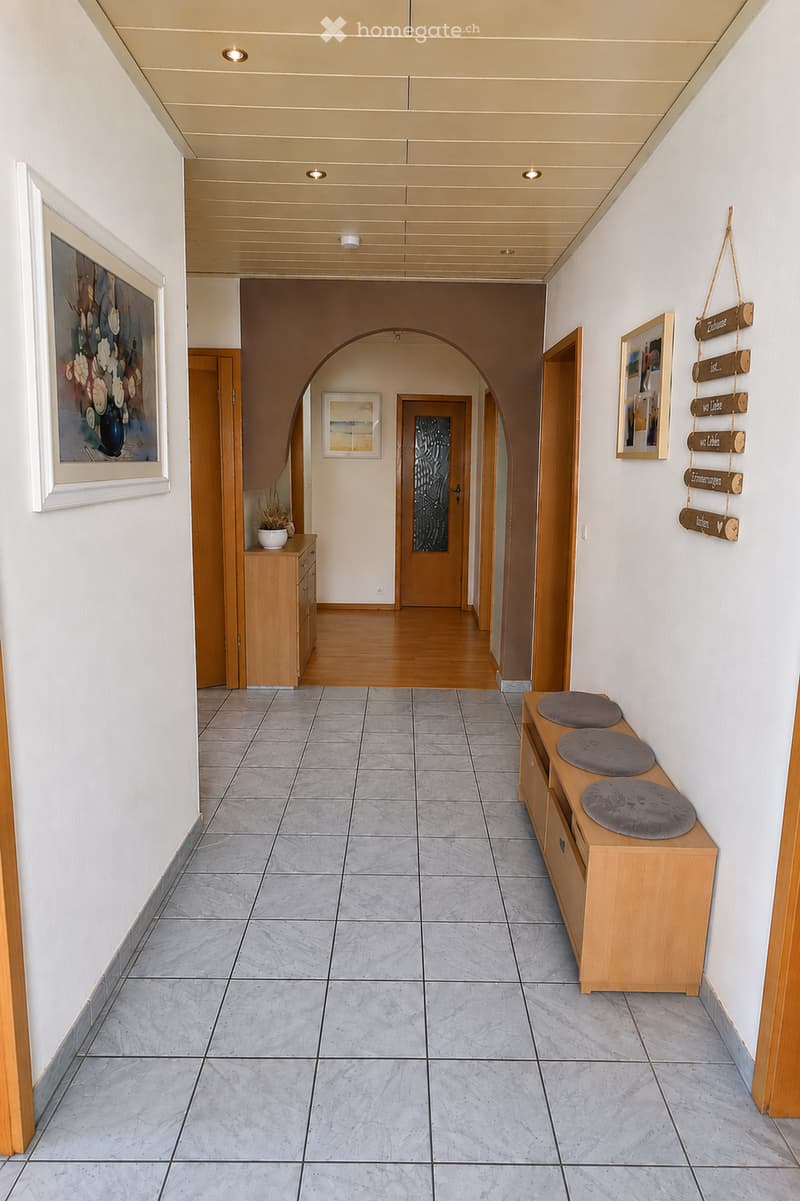 Spacious hallway with tiled floor, wooden ceiling, and several doors leading to different rooms. Features include a small wooden cabinet with a plant, a bench with cushions, and decorative wall signs.
