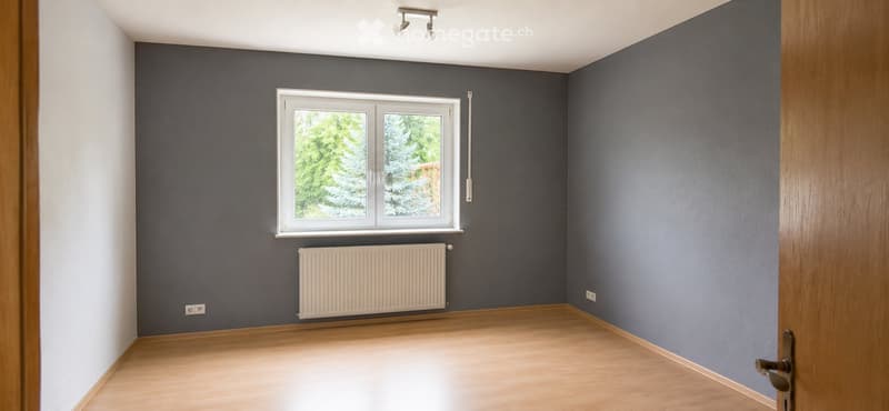 Empty room with wooden flooring, grey walls, a window with white frame, radiator, and a door with a brass handle.