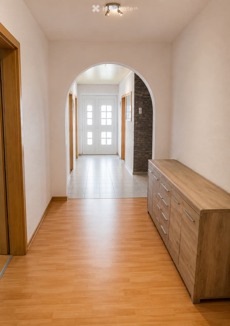 Empty hallway with wooden flooring, light pink walls, and a wooden sideboard on the right. An arched passage leads to another area with white doors and tiled flooring.