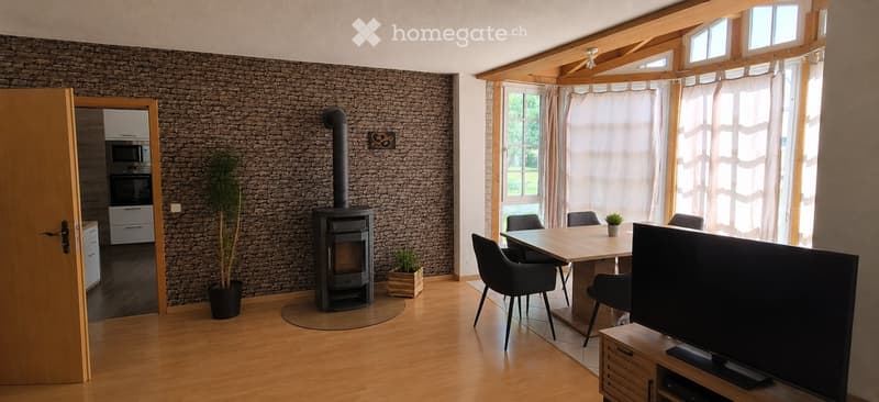 Cozy living room with wooden flooring, exposed wooden beams, large windows with curtains, fireplace, dining table with chairs, and television.