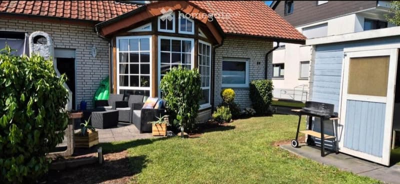 Garden area with a patio, outdoor seating, and a barbecue. The house has a red-tiled roof and large windows.