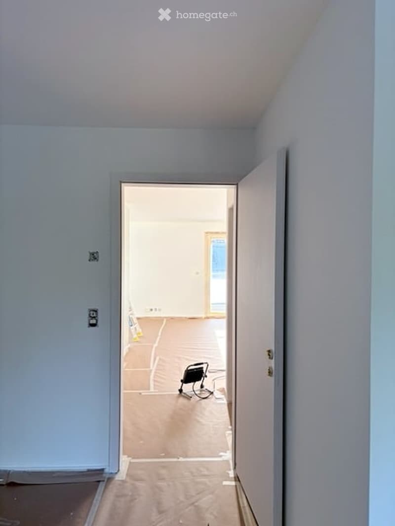 Empty room with light blue walls and a wooden floor, showing an open doorway leading to another room with a visible floor lamp.
