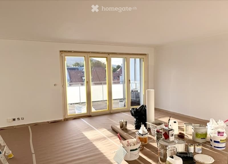 Empty room with wooden flooring, white walls, large sliding glass doors leading to a balcony, and construction materials scattered on the floor.