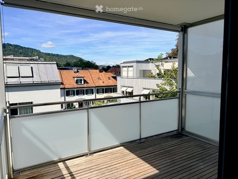 Balcony with wooden decking, glass railing, and scenic view of surrounding buildings and hills.