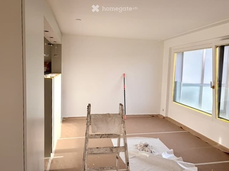 Empty room with white walls, wooden floor, and large windows. Ladder and cleaning supplies are present, indicating ongoing renovation.
