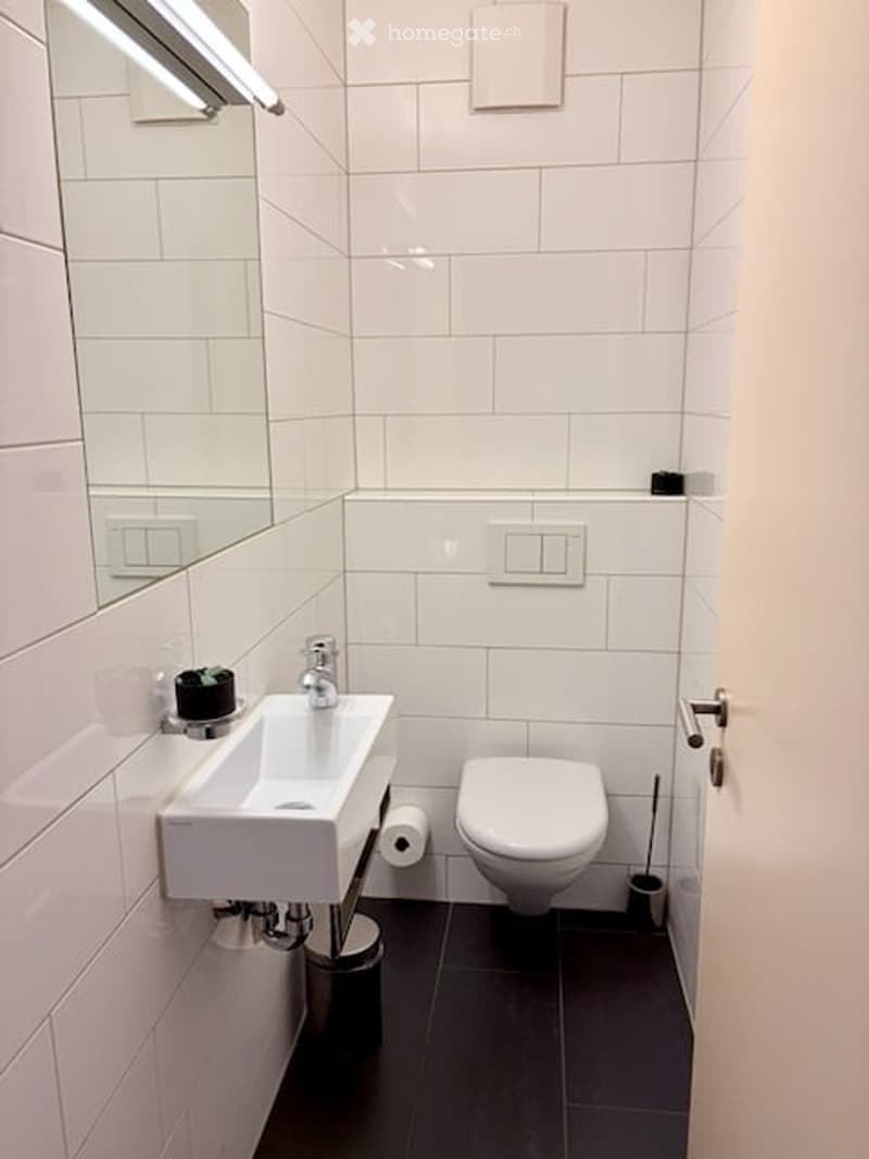 Modern, compact bathroom with white tiles, a white sink, a toilet, a mirror, and a cup on the sink counter.