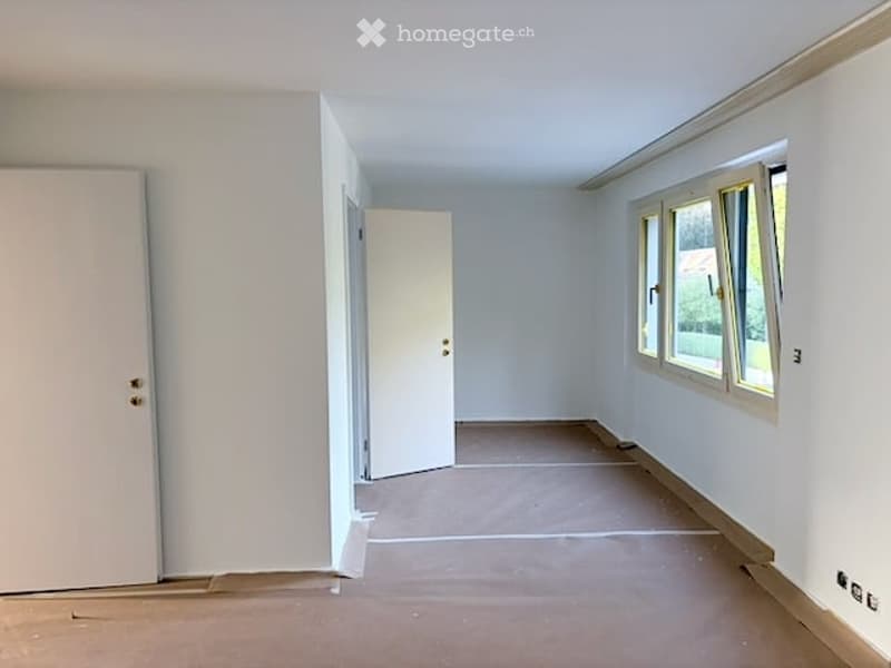 Empty room with white walls, wooden floor, and two doors. Windows on one side allow natural light.