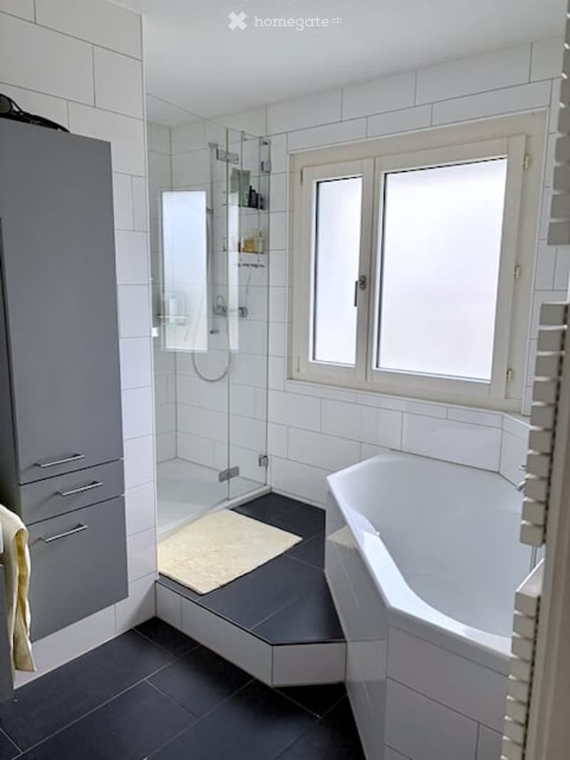 Modern bathroom with a white bathtub, glass-enclosed shower, grey cabinet, and a large window providing natural light.