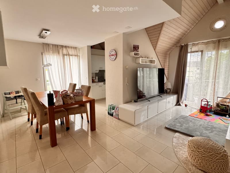 A modern, open-plan living and dining area with a large flat-screen TV, a dining table with chairs, a woven stool, and a small kitchen area visible in the background.
