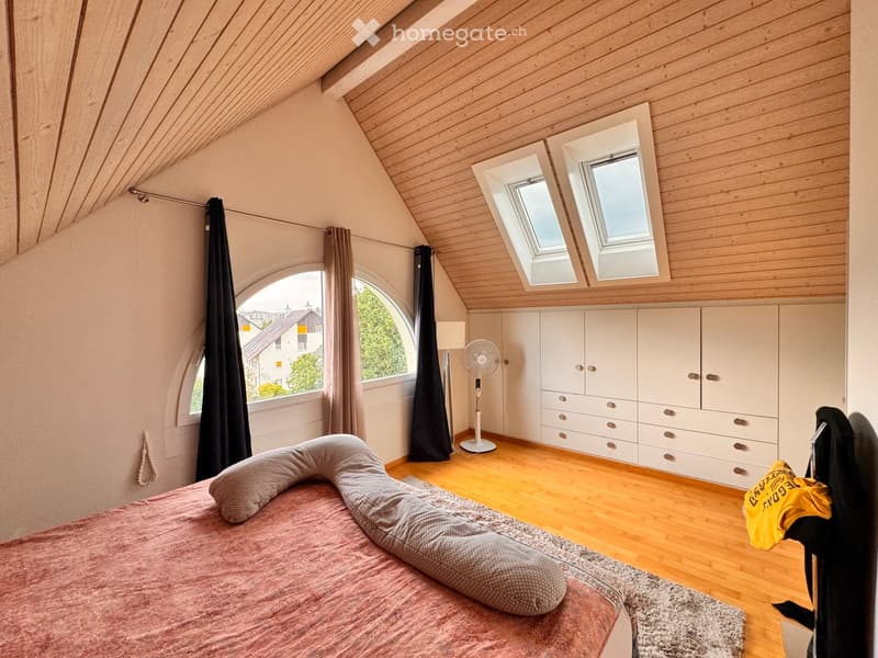 Cozy attic bedroom with wooden slanted ceilings, large windows, and built-in white wardrobes. Features a pink bed with pillows, a fan, and a small rug.