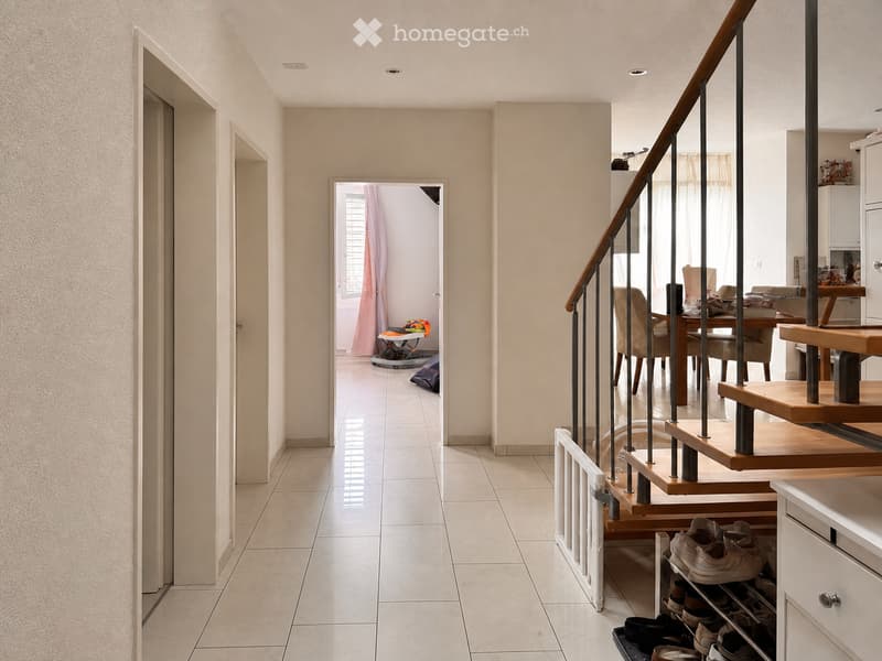 A well-lit hallway with tiled flooring, a staircase with wooden steps and metal railings, and a shoe rack with several pairs of shoes. The hallway leads to an open area with a dining table and chairs visible in the background.
