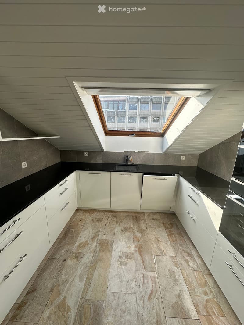 Attic kitchen with white cabinets, black countertops, and a skylight window providing natural light. The floor is covered with beige tile.