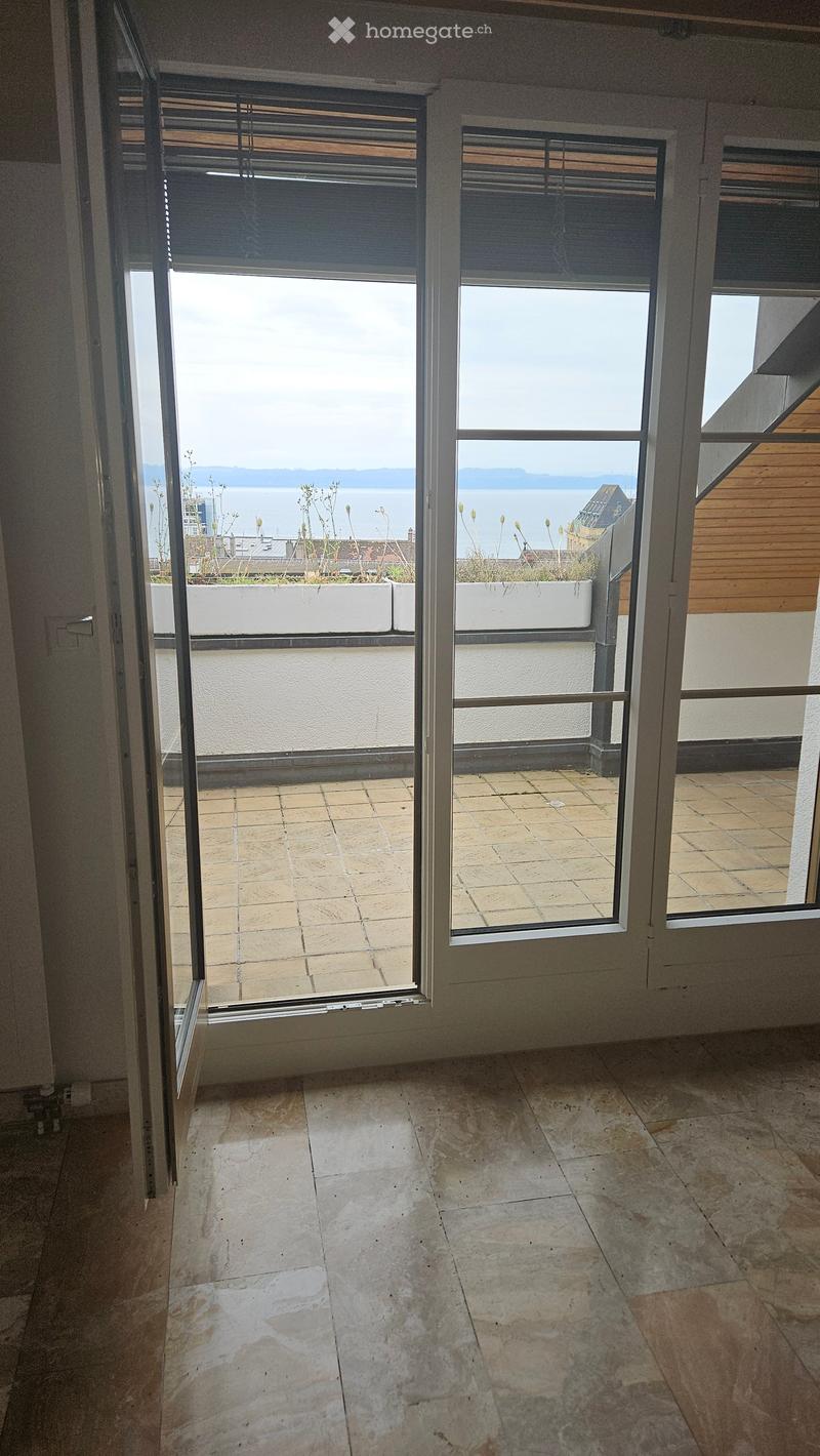 The image shows a room with large windows that provide a view of the outdoors. The floor is covered in tile, and there are blinds on the windows. The room appears to have access to a balcony or terrace area.