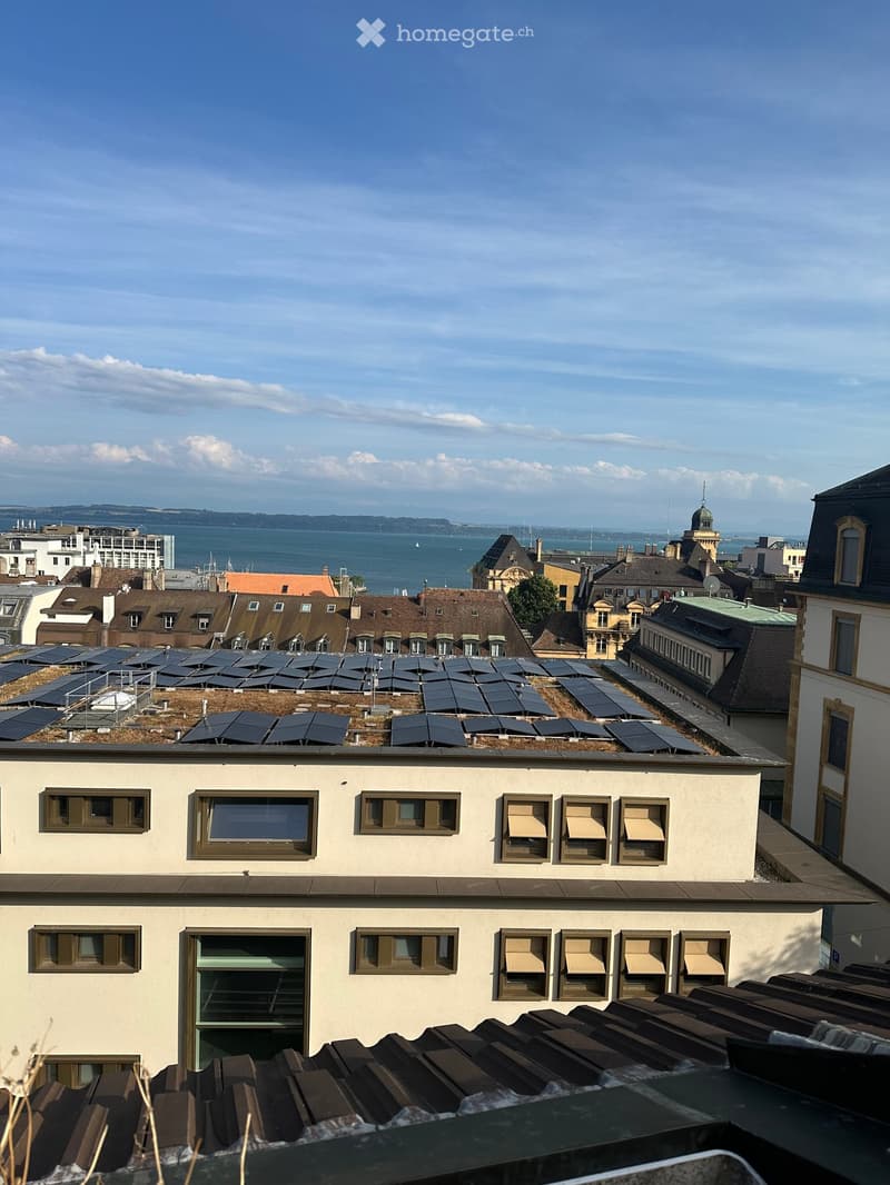 The image shows a cityscape with a view of the harbor and water in the background. The foreground features a multi-story building with a slanted roof and several windows. On the roof, there are solar panels installed, indicating the use of renewable energ