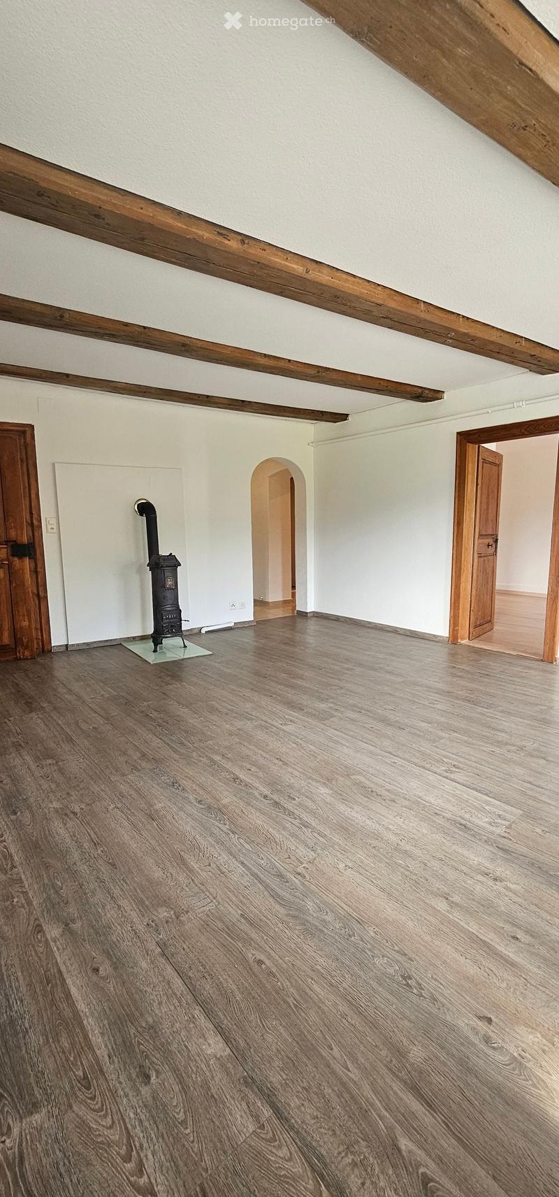 Empty room with wooden floor, white walls, exposed wooden beams, and a small black stove with a chimney. There are two wooden doors and an arched doorway leading to another room.