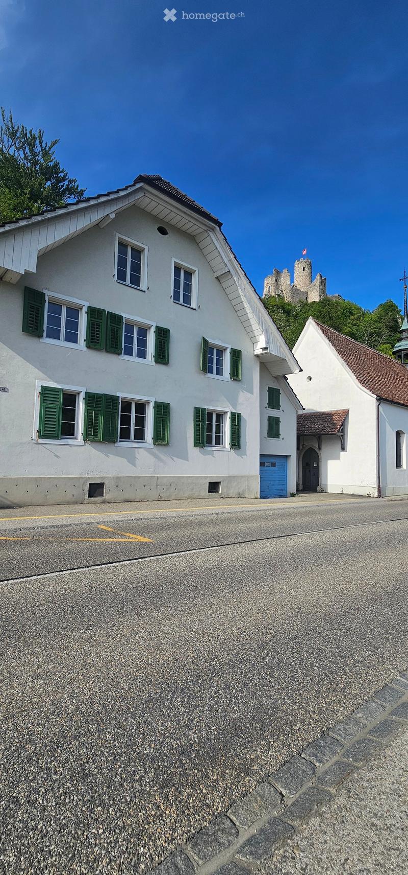 A two-story white building with green shutters and a blue garage door, situated on a raised ground floor with a stone retaining wall. A small church with a red-tiled roof is visible to the right, and a castle-like structure is seen in the background on a
