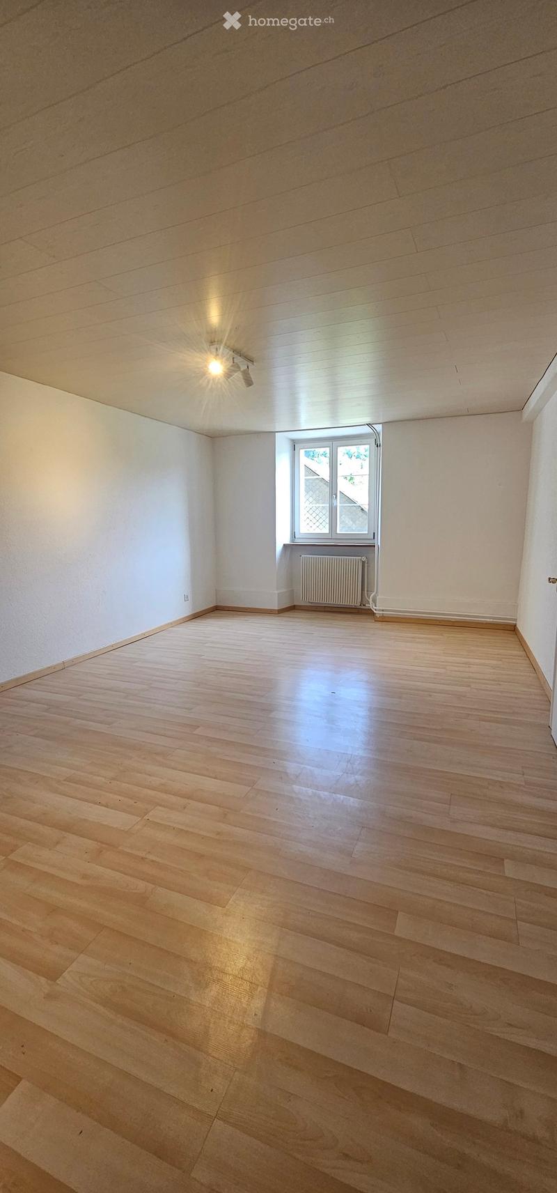Empty room with light wooden flooring, white walls, and a single window with a radiator below it.