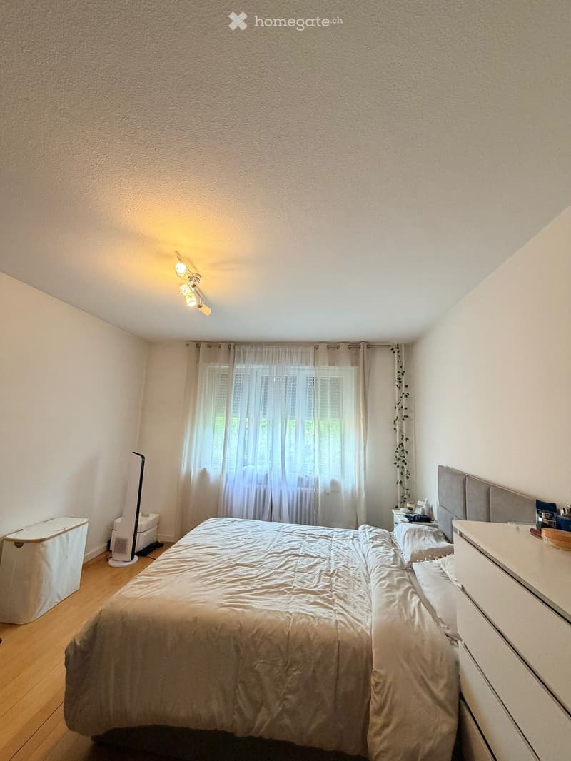A well-lit bedroom with a large bed, nightstand, fan, and window with curtains.
