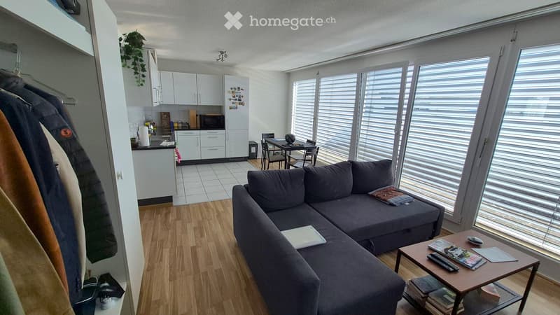 A modern, open-concept living and dining area with a grey sectional sofa, coffee table, dining table with chairs, and a small kitchen area with white cabinets and a refrigerator.