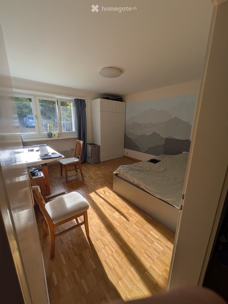 Bright bedroom with wooden flooring, a bed with a mountain-themed headboard, a small table with chairs, and a cabinet.