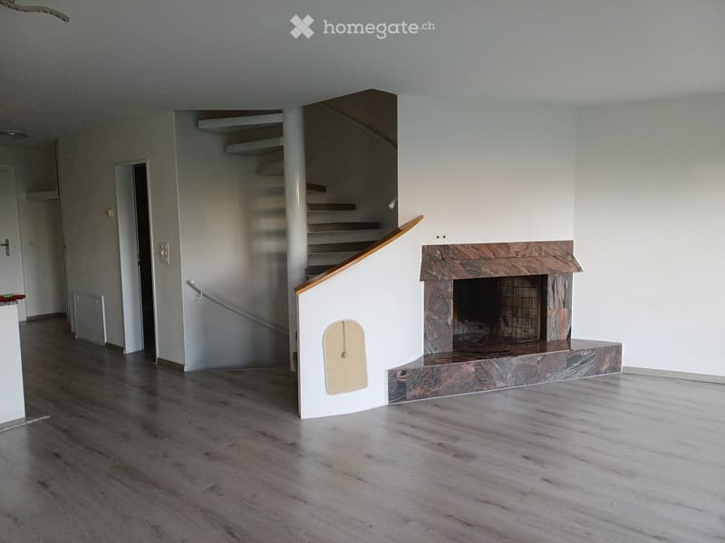 Empty room with wooden flooring, white walls, and a stone fireplace. A staircase leads to an upper level, and a small pet door is visible on the wall.