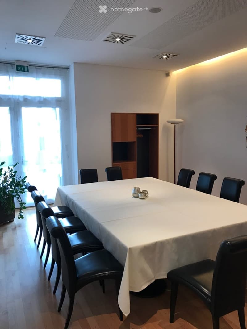 A conference room with a long table covered in white cloth. Black chairs are arranged around the table. The room features a wooden cabinet with glass doors. A potted plant and floor lamp are in the corner. Windows on the wall allow natural light.