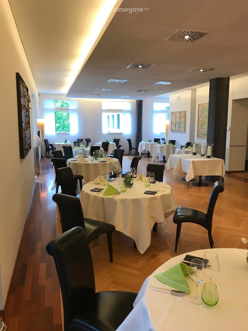 Empty restaurant with multiple tables covered with white cloths, black chairs around, wooden floor, paintings on the wall, windows with shades, lamps on the wall