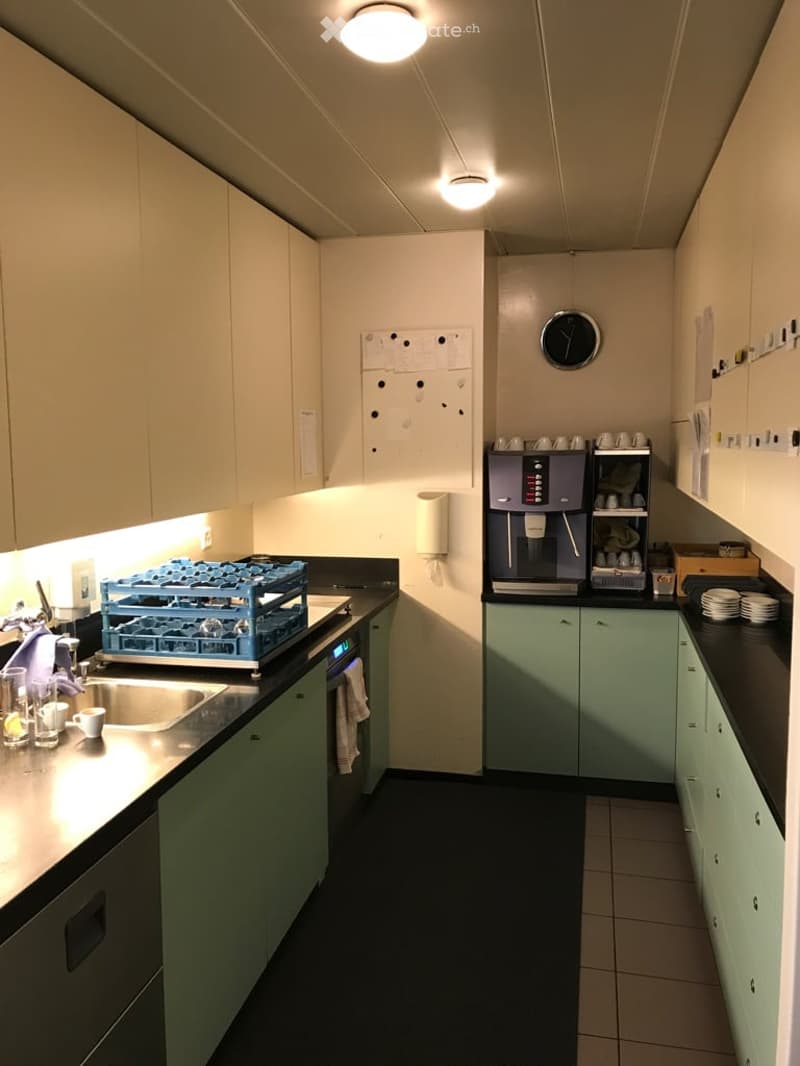 A kitchen with a sink, cabinets, a coffee machine, countertop, shelves, and a clock.