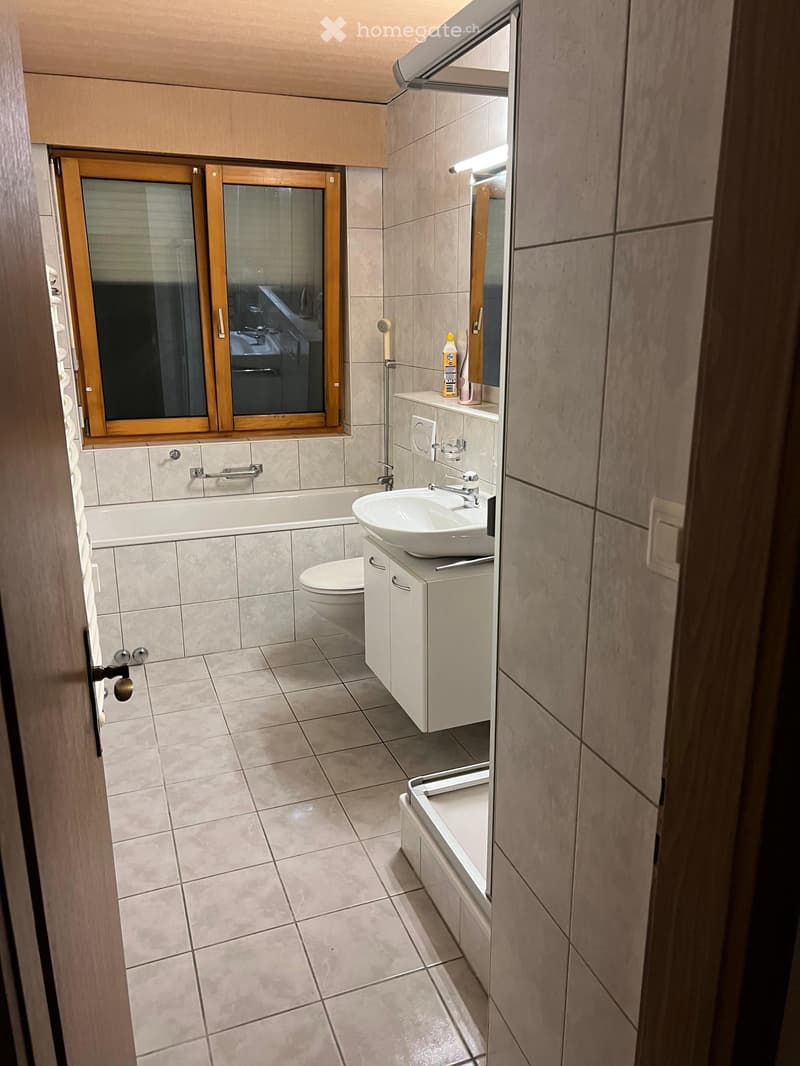 Modern bathroom with tiled walls and floor, featuring a bathtub, sink, toilet, and a cabinet with a mirror above it. A window with wooden frame is visible above the bathtub.