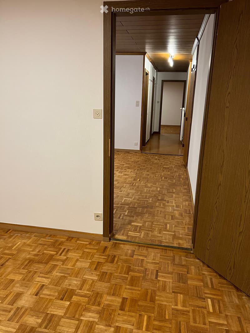 Empty hallway with wooden herringbone flooring, white walls, and recessed lighting. Doors are visible along the corridor.