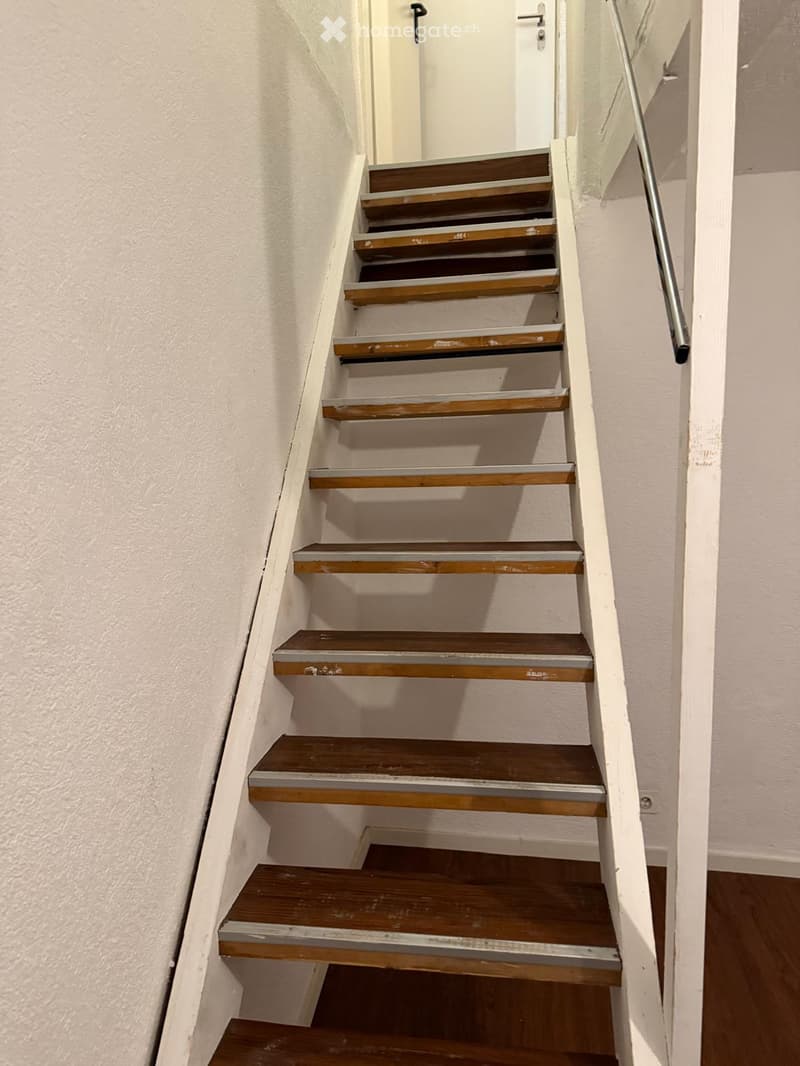 Wooden staircase with white walls and a metal handrail, leading to an open doorway at the top.
